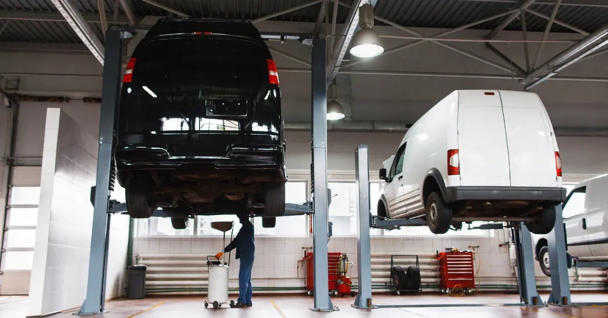 Auto service bay with two gray two‑post lifts raising a black van and a white van while a technician works underneath the black van using an oil drain cart