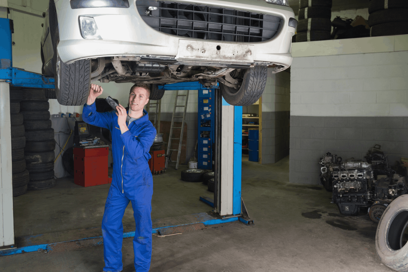 Auto mechanic inspecting the underside of a car raised on a two-post lift in a repair shop.