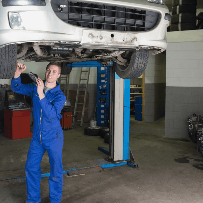 Auto mechanic inspecting the underside of a car raised on a two-post lift in a repair shop.