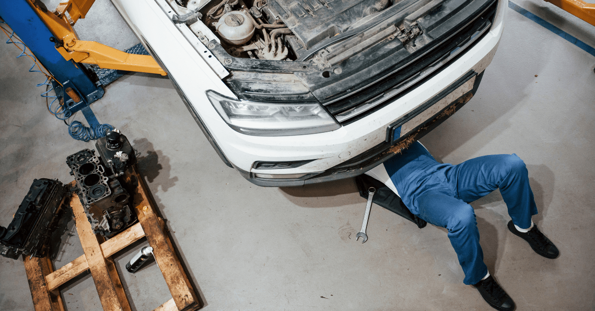 Auto mechanic performing undercar maintenance on a lifted vehicle with tools and engine parts nearby.