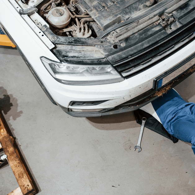 Auto mechanic performing undercar maintenance on a lifted vehicle with tools and engine parts nearby.