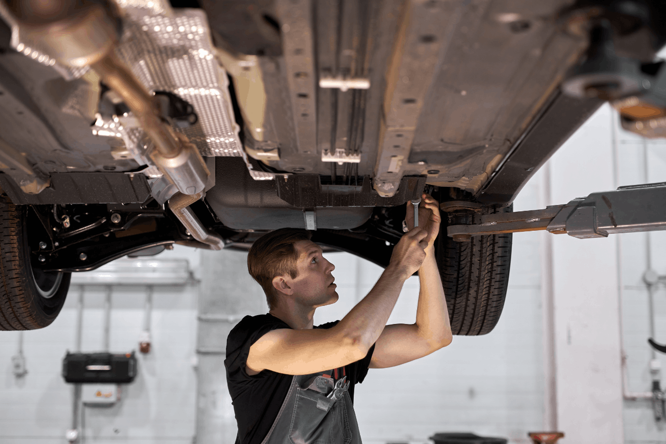 Auto mechanic using a wrench beneath a vehicle raised on a two-post lift in a repair shop.