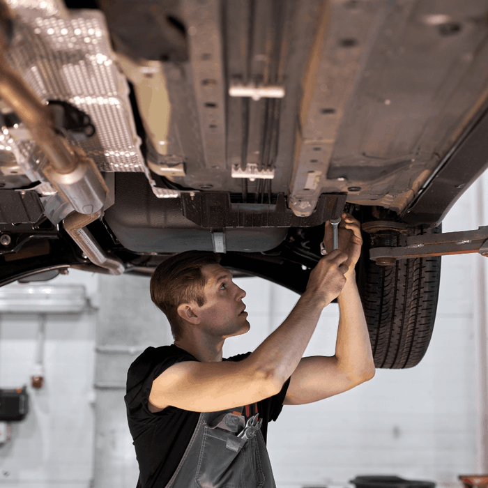 Auto mechanic using a wrench beneath a vehicle raised on a two-post lift in a repair shop.