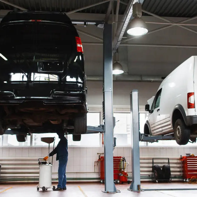 Auto service bay with two gray two‑post lifts raising a black van and a white van while a technician works underneath the black van using an oil drain cart