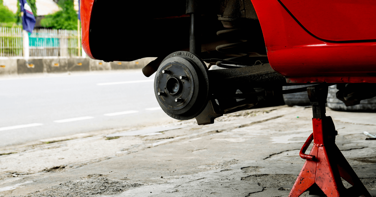 Car on jack stands with wheel removed, drum brake and suspension visible on driveway.