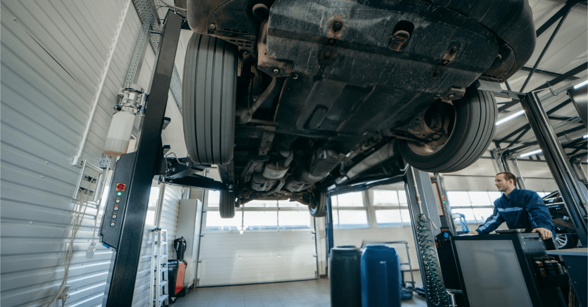 Mechanic operating a 2-post car lift in an auto repair shop with a vehicle raised for undercarriage inspection.