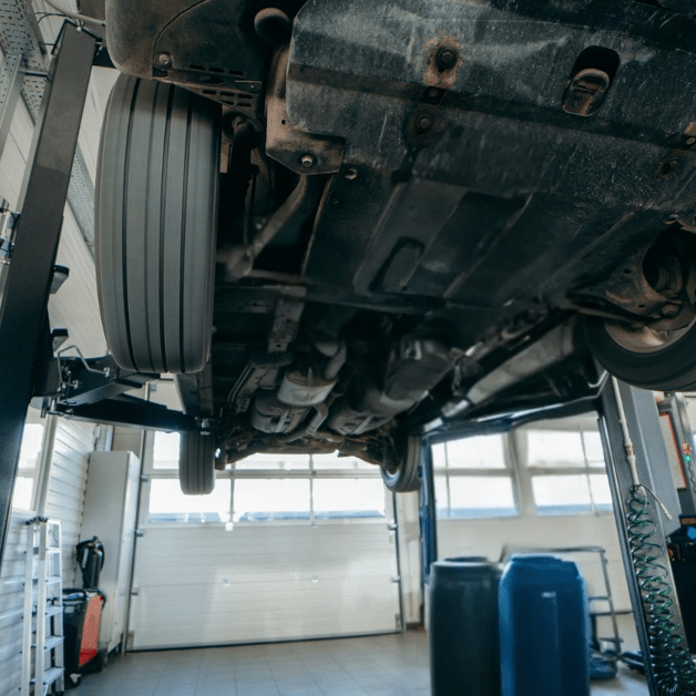 Mechanic operating a 2-post car lift in an auto repair shop with a vehicle raised for undercarriage inspection.