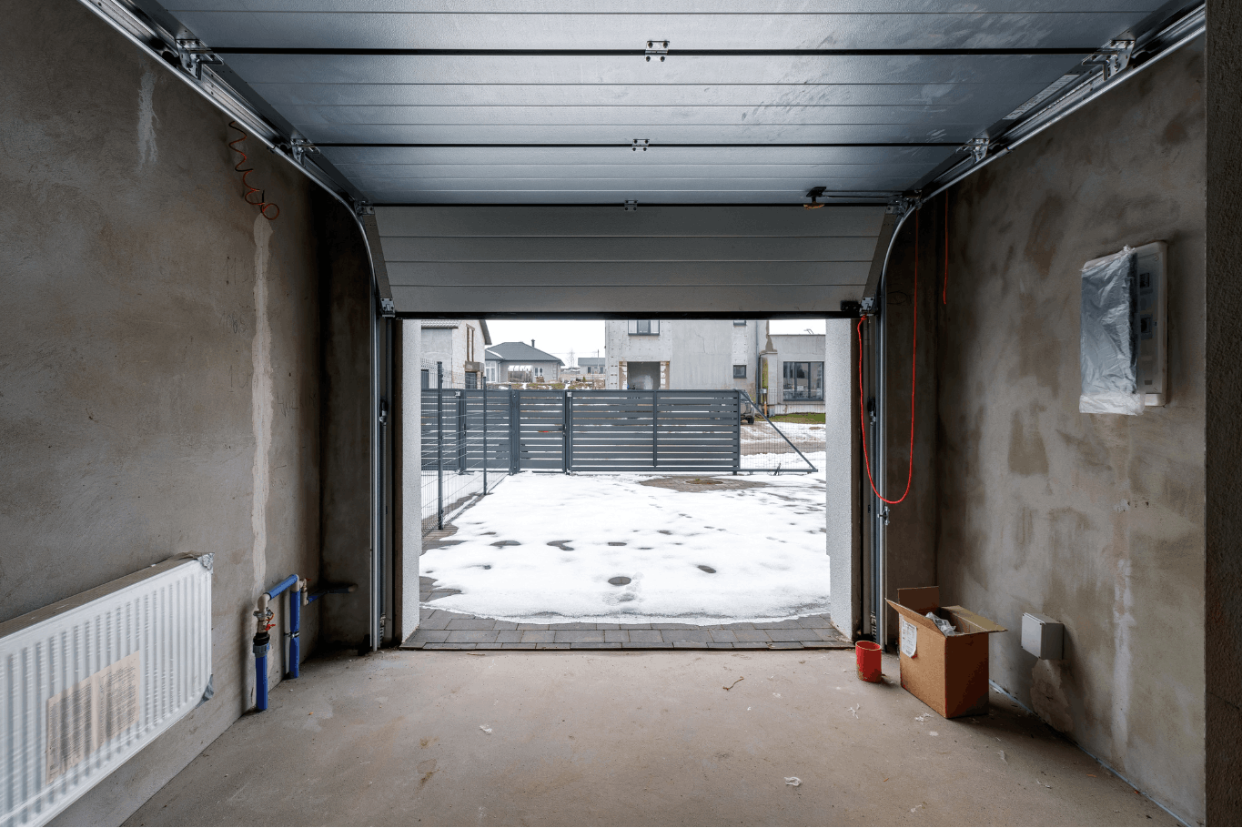 Partially open sectional garage door with unfinished garage interior and snowy driveway outside