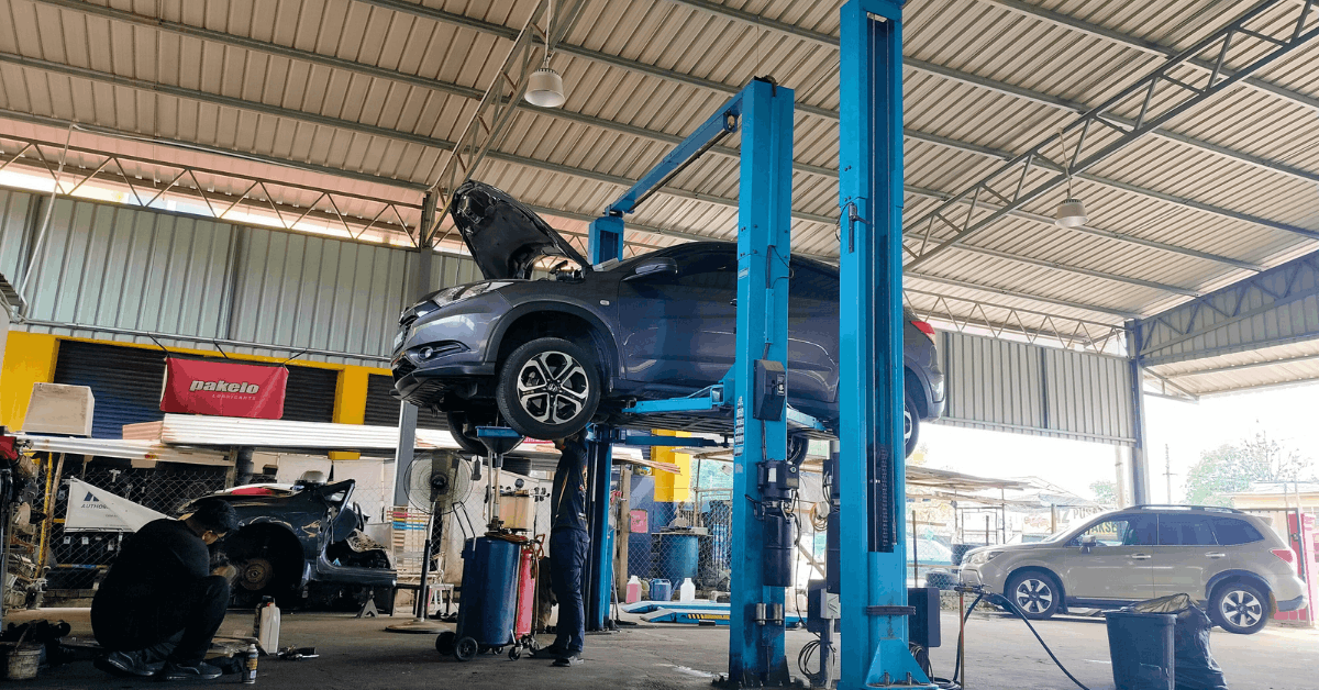 Vehicle lifted on a two post automotive lift in a service bay with the hood open and a mechanic performing maintenance below
