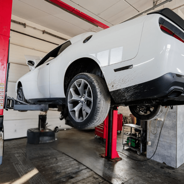 White Dodge Challenger lifted on a red 2-post car lift inside an auto repair shop.