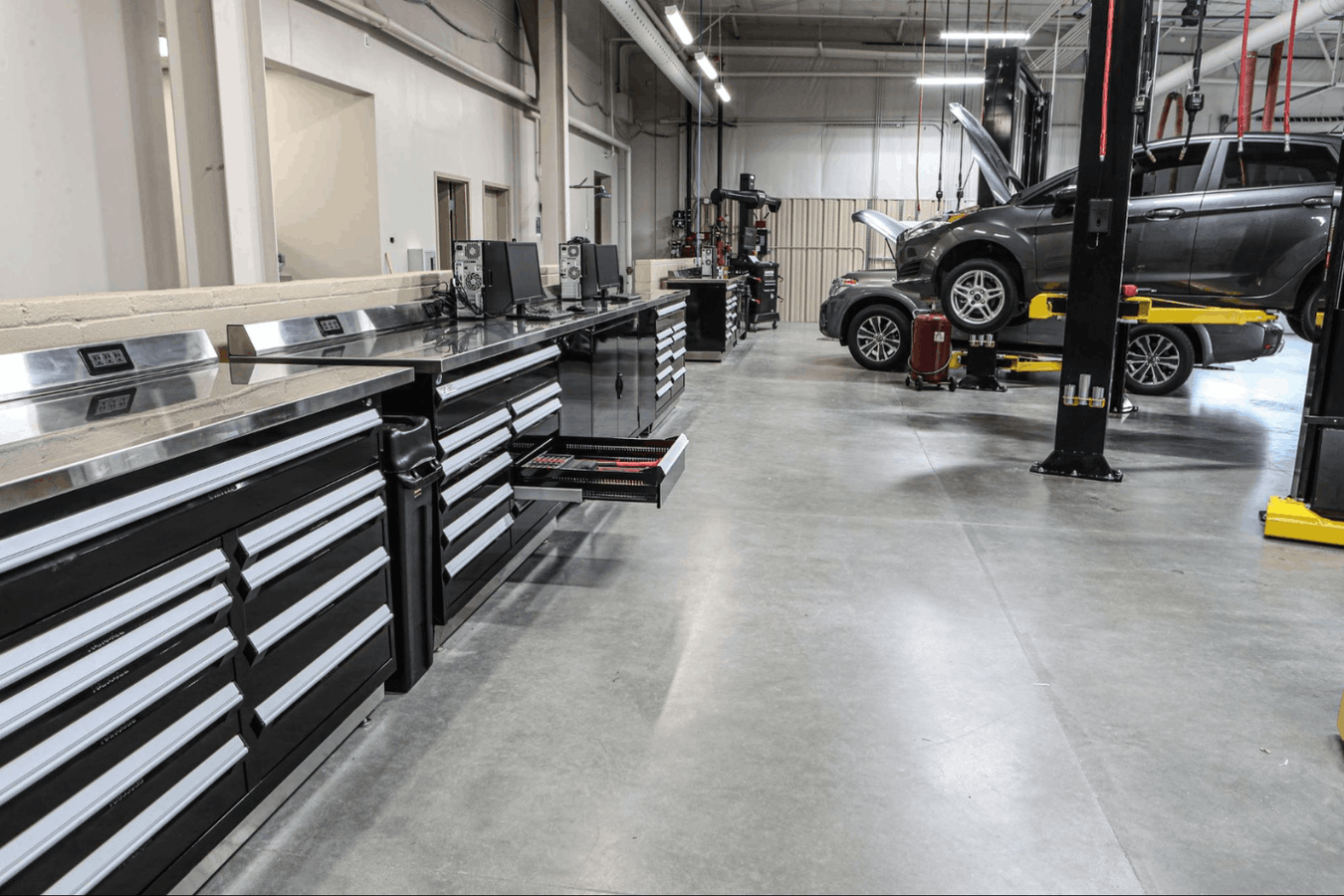 Commercial garage workbench section showing long stainless workbenches with drawer cabinets and a vehicle raised on a two post car lift in the background